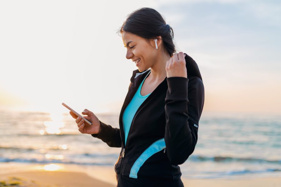 young attractive slim woman doing sport exercises on morning sunrise beach in sports wear, healthy lifestyle, listening to music on wireless earphones holding smartphone, smiling happy having fun